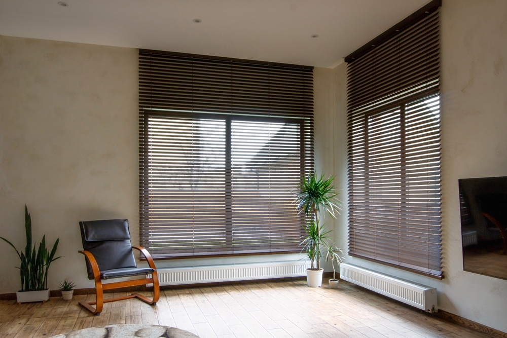 Wooden blinds on large windows in the interior. Living room with armchair and houseplants near windows with wood blinds.