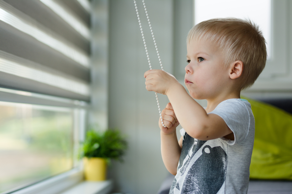 Little, three years old boy he raises the blinds in the windows and looking out the window.